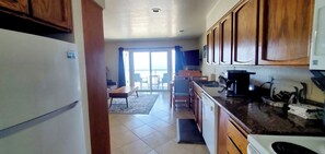 A kitchen with wooden cabinets, a refrigerator, a coffee maker, and a dining area with a table and chairs. - Condo #108 with beautiful sunset view from your balcony overlooking the ocean (Lincoln City)
