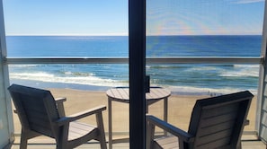 Un balcon avec deux chaises et une petite table, donnant sur une plage où l’on peut admirer les vagues et un ciel dégagé.