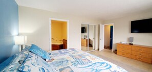 A bedroom with a bed, a dresser, a television, and a bathroom visible through an open door. - Condo #307 with beautiful sunset view from your balcony overlooking the ocean (Lincoln City)
