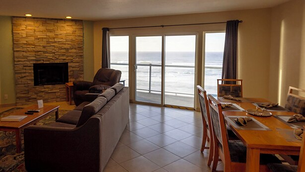 A living room with a stone wall, a flat-screen TV, a sofa, a chair, a dining table with chairs, and a view of the ocean through large glass doors. - Condo #307 with beautiful sunset view from your balcony overlooking the ocean (Lincoln City)