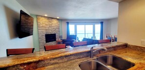 A modern living room with a stone wall, a flat-screen TV, and a large window overlooking the ocean. - Condo #307 with beautiful sunset view from your balcony overlooking the ocean (Lincoln City)