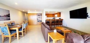 A living room with a sofa, dining table, chairs, and a kitchen area with wooden cabinets and a TV mounted on the wall. - Condo #307 with beautiful sunset view from your balcony overlooking the ocean (Lincoln City)