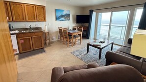 A living room with a sofa, a dining table with chairs, a kitchen area with wooden cabinets, a television, and a large window with a view of the ocean. - Condo #206 with beautiful sunset view from your balcony overlooking the ocean (Lincoln City)
