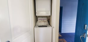 A compact, white laundry room with a built-in washer and dryer, a door leading to a hallway, and a blue door visible in the background. - Condo #203 with beautiful sunset view from your balcony overlooking the ocean (Lincoln City)
