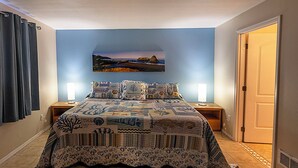 A bedroom with a large bed, two bedside tables, a nightstand, a wall-mounted painting, a door, and a patterned rug. - Condo #207 with beautiful sunset view from your balcony overlooking the ocean (Lincoln City)