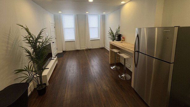 A compact kitchen with a stainless steel refrigerator, wooden bar stools, and two potted plants.