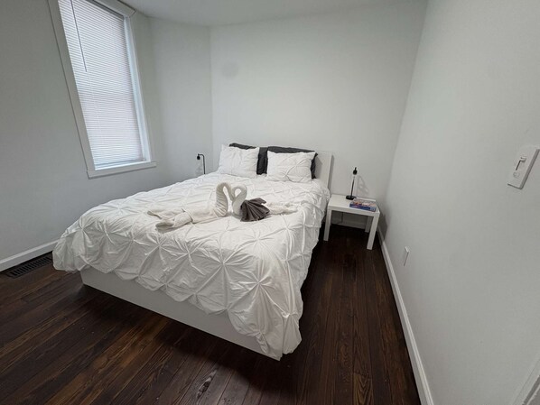 A neatly made bed with a white duvet and decorative swan pillows, a bedside table with a lamp, and a window with blinds. - Temple Apartment Unit 1 2 bedrooms (Philadelphia)
