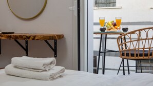 Une chambre d’hôtel avec un lit, une table avec une télécommande, un fauteuil en osier et un balcon avec des éléments pour le petit-déjeuner.