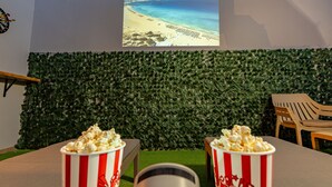 Une pièce avec un grand écran diffusant une scène de plage, deux seaux de pop-corn posés sur une table et un mur recouvert de plantes vertes.