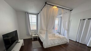 A bedroom with a four-poster canopy bed, a flat-screen TV, a bedside table, and a window with curtains. - Temple Apartment Unit 2 1 bedroom (Philadelphia)