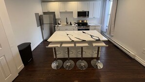 A modern kitchen with white cabinets, a central island with bar stools, stainless appliances, and a window with blinds. - Temple Apartment Unit 2 1 bedroom (Philadelphia)