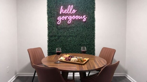 A dining area with a round wooden table, four brown chairs, a neon sign on the wall, and a fruit platter with wine glasses. - Music Weekend in Woodstock| Madlife|Lamplighter|Am (Woodstock)