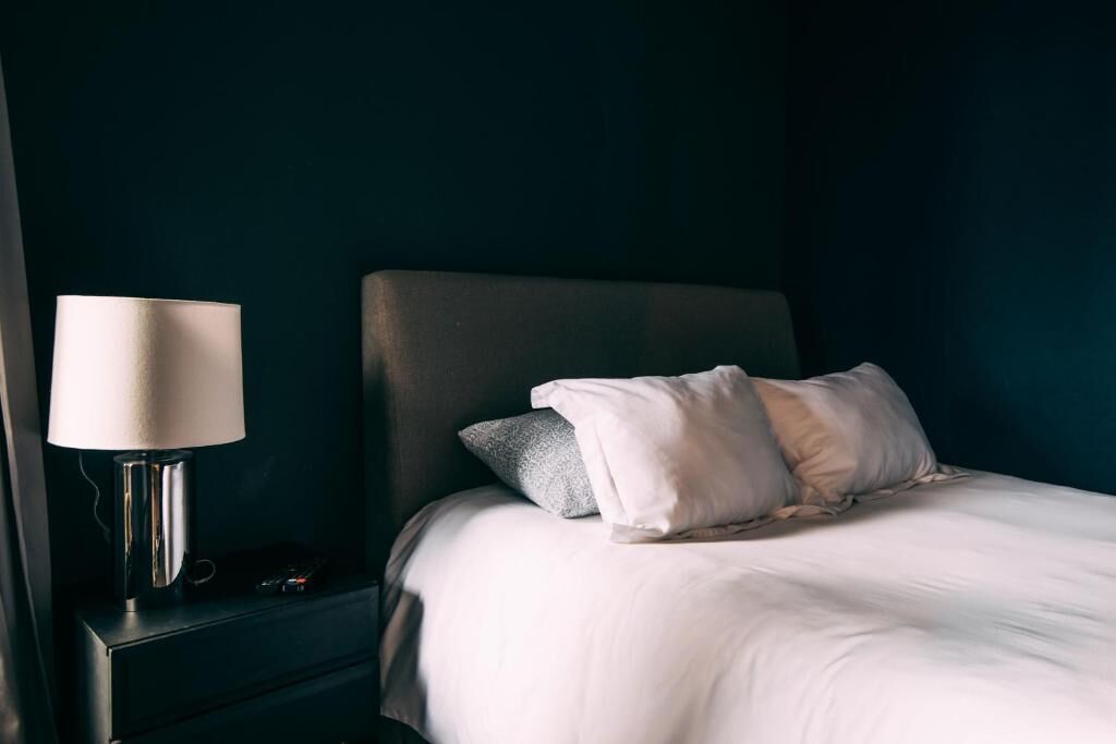 A neatly made bed with white bedding and a grey pillow, a bedside lamp, and a dark-colored nightstand.