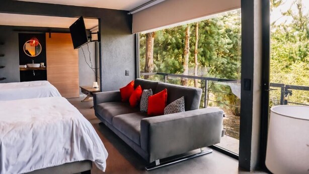 Ein modernes Schlafzimmer mit einem Bett, einem grauen Sofa mit roten und gemusterten Kissen, einem an der Wand montierten Fernseher und einem großen Fenster mit Blick auf Bäume.