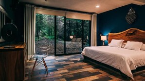 Ein Schlafzimmer mit einem großen Bett, einem Schreibtisch und einem Stuhl. Durch eine Glastür ist ein Blick auf einen Wald möglich.