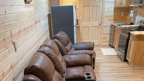 Un salon chaleureux avec un canapé en cuir marron, des murs recouverts de panneaux en bois, un coin cuisine doté de meubles en bois et un réfrigérateur.