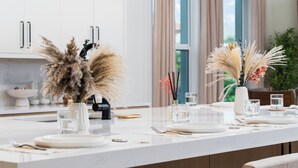 A modern kitchen with white cabinets, a marble countertop, and a dining area set with white plates, glasses, and a vase with dried flowers. - Ocean Hammock Luxury with Pool, Game Room & Beach Access (Palm Coast)