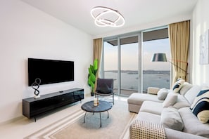 Un salon moderne doté d’un téléviseur à écran plat, d’un meuble de rangement noir, d’une table basse ronde, d’un canapé gris et d’une grande fenêtre offrant une vue sur la mer.