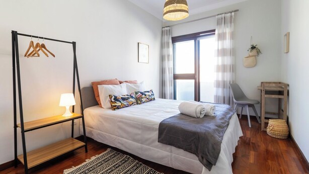 A neatly arranged bedroom with a bed, a bedside table with a lamp, a chair, a small table, and a hanging plant. - Porto Essence Apartment (Porto)