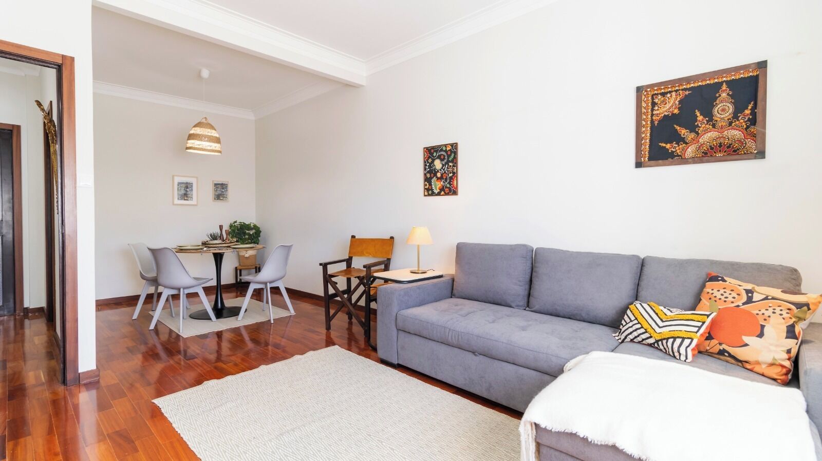 Een moderne woonkamer met een grijze bank, een eettafel met stoelen, een klein bijzettafeltje met een lamp en een decoratieve wandversiering.