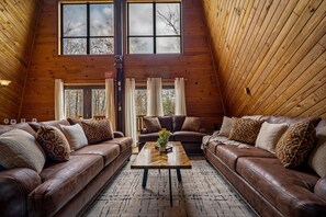 A cozy loft-style room with two leather sofas, a wooden coffee table, and a large window with curtains. - New Listing! 18ft Swim Spa, Huge Game Rm, Central Location! (Gatlinburg)