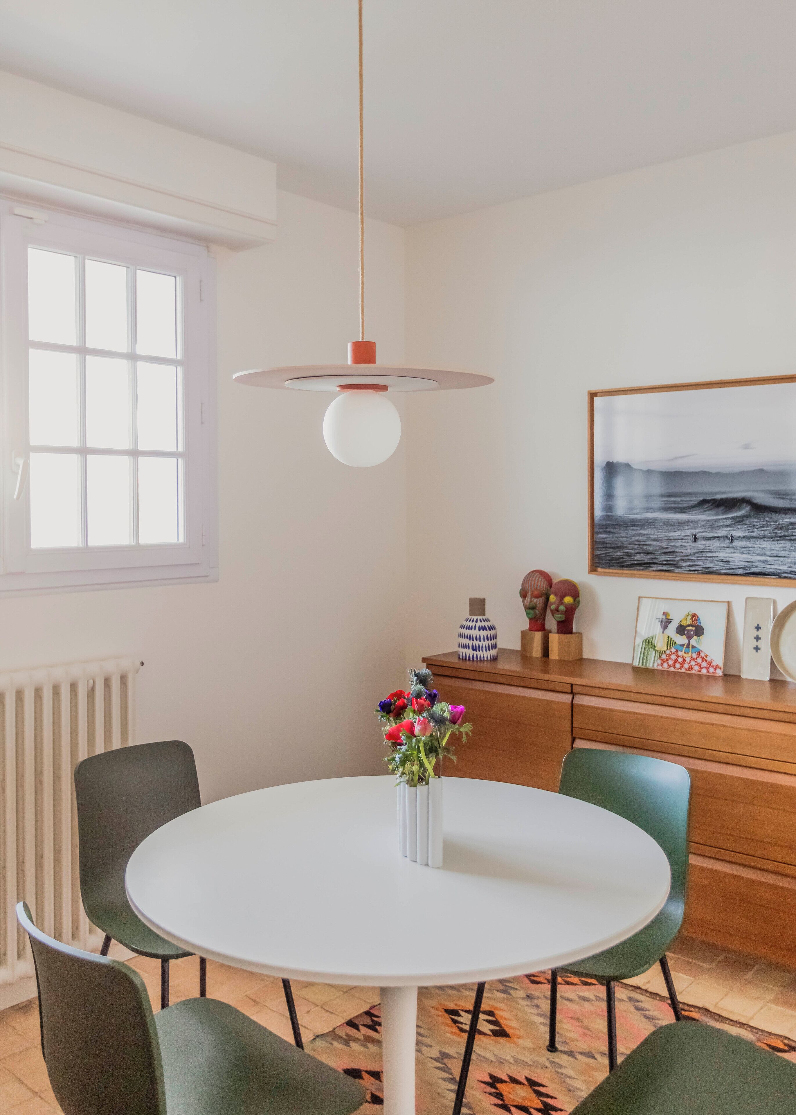 Un coin repas avec une table ronde blanche, des chaises vertes, un luminaire suspendu, un vase avec des fleurs et un meuble en bois orné d’objets décoratifs.