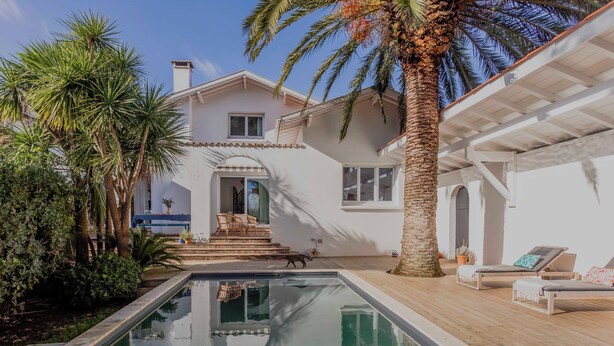 A poolside area with a clear pool, a covered seating area, and a house with a balcony.