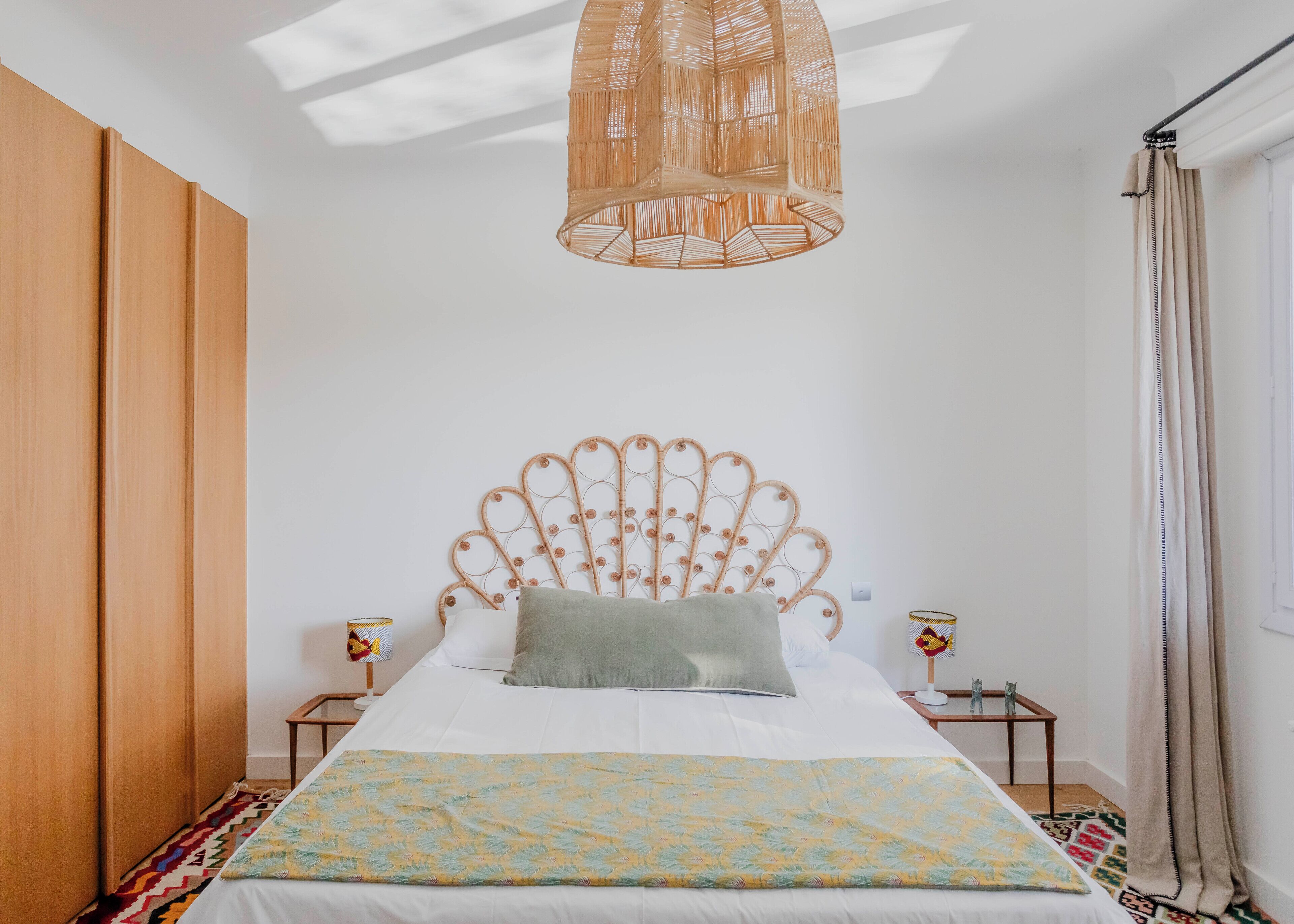 A bedroom with a bed, a bedside table, a nightstand, a hanging light fixture, and a window with curtains.