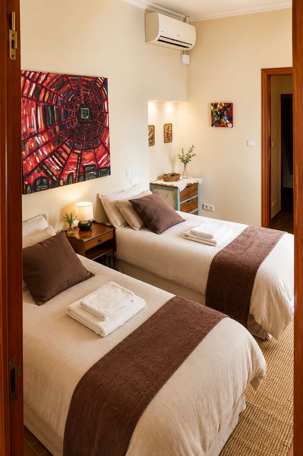 Habitación de hotel con dos camas, aire acondicionado instalado en la pared, mesita de noche con lámpara y obras de arte en las paredes.