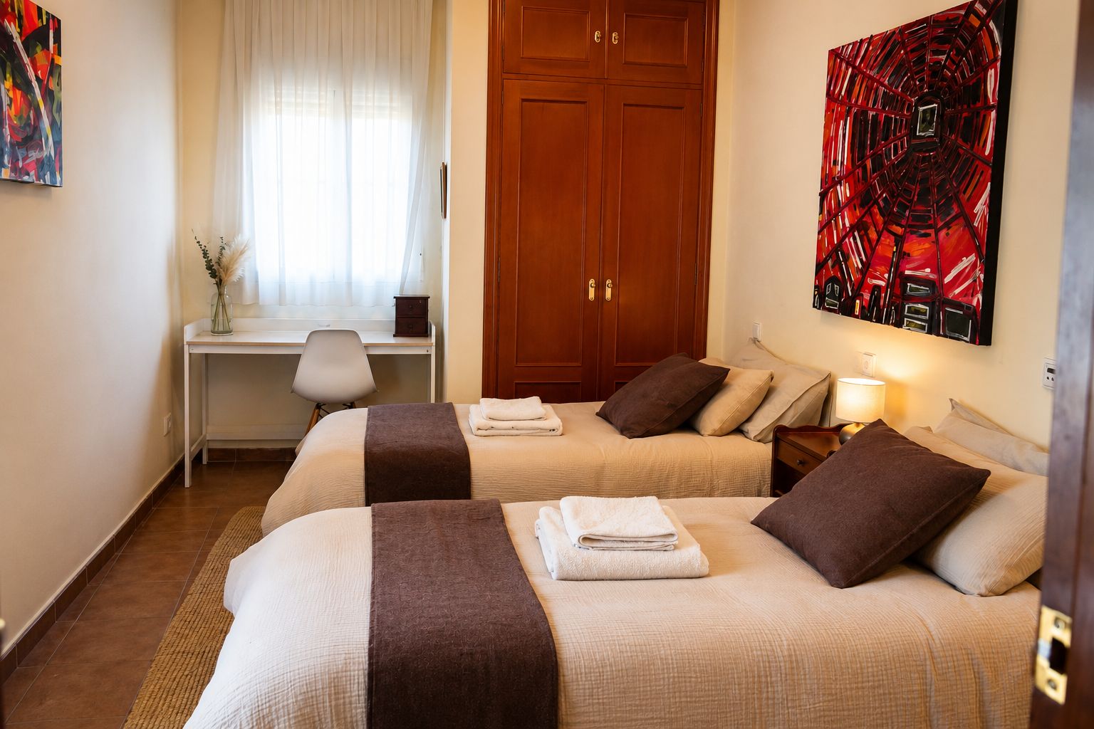 Habitación de hotel con dos camas, un escritorio, una silla, una ventana con cortinas y una obra de arte en la pared.