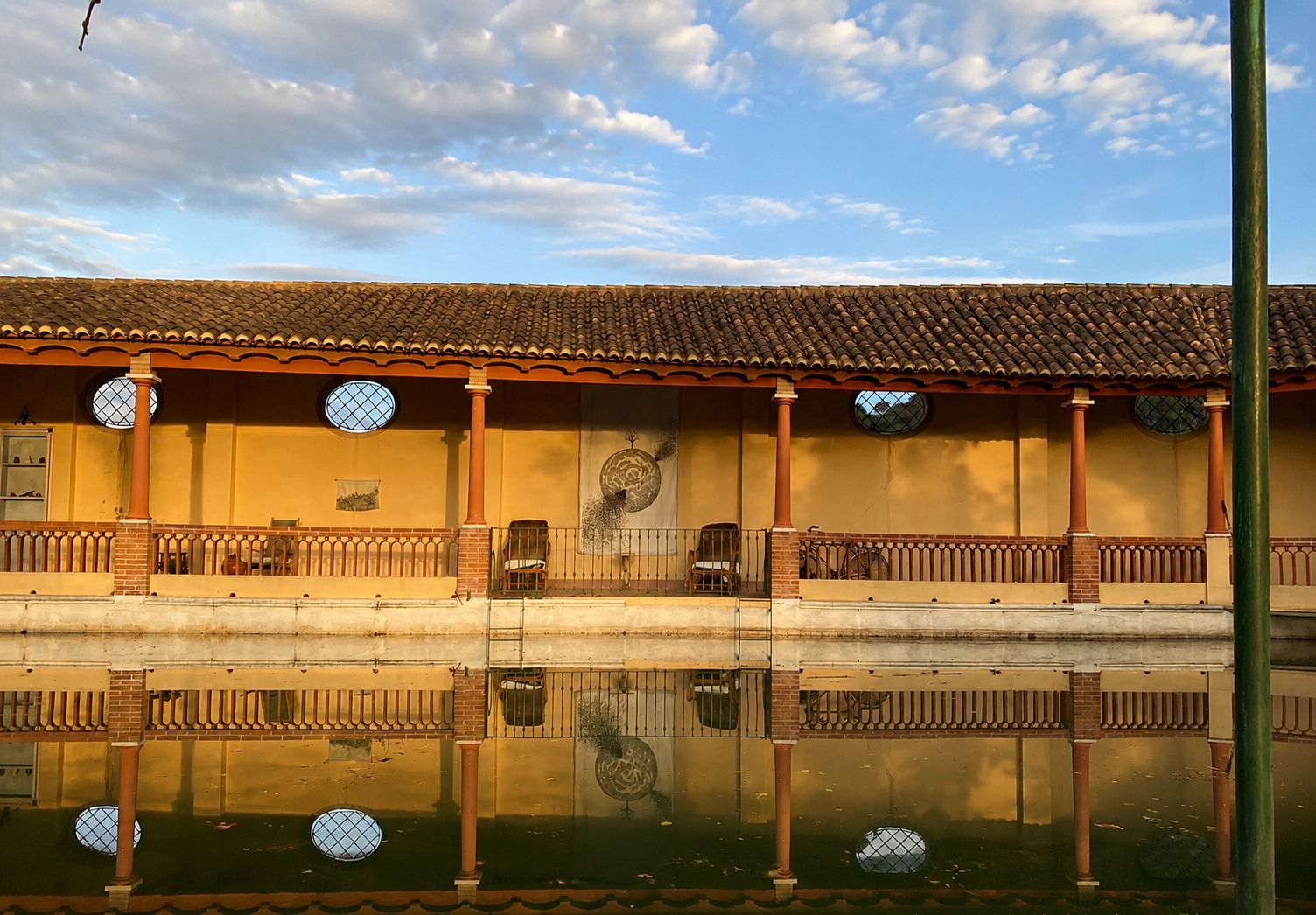 A building with a tiled roof, a balcony with a railing, and a reflection in the water.