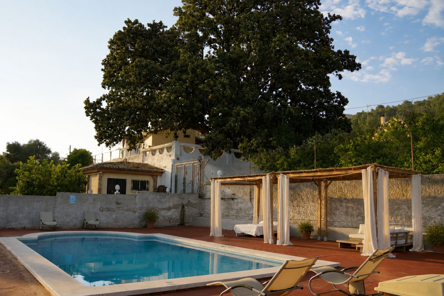 Una piscina con sillas de descanso, una pérgola con cortinas y un edificio con tejado de baldosas.