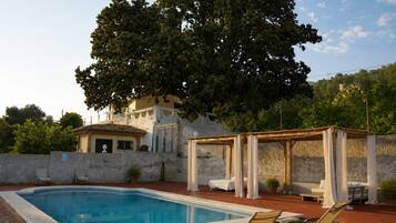 Una piscina con sillas de descanso, una pérgola con cortinas y un edificio con tejado de baldosas.