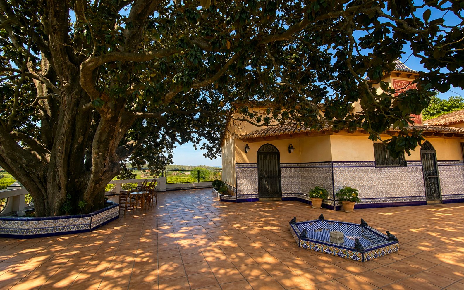 Un patio con un árbol grande, suelo de baldosas, un edificio con fachada de azulejos y una pequeña zona de estar.