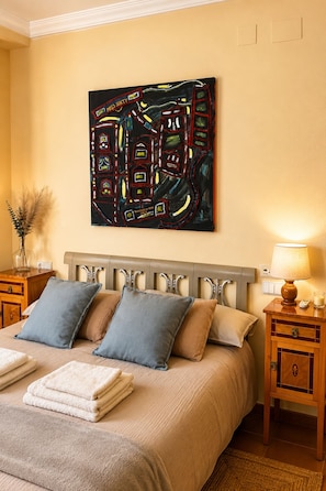 A hotel room with a bed, pillows, a bedside table with a lamp, a wall-mounted abstract painting, and a vase with dried plants. - Historic Villa with Private Pool & Gardens near Valencia (Alzira)