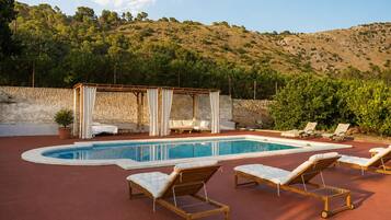 Zona de piscina con sillones de descanso, una pérgola y vistas a una colina.