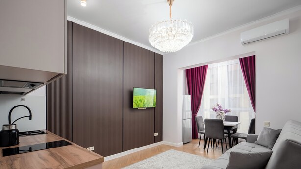 A modern living room with a grey sofa, a dining area with a table and chairs, a kitchen with a stove and sink, and a large window with curtains.