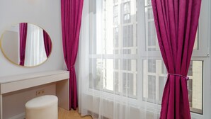 A room with a white desk, a round mirror, a white ottoman, and a window with sheer white curtains and a purple tieback.
