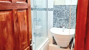 Un baño con puerta de madera, cabina de ducha de vidrio, lavabo blanco y salpicadero de azulejos.