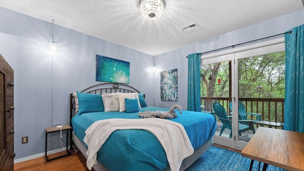 A bedroom with a bed, a small table, a chair, a balcony with a view, and a ceiling light. - Pretty Peacock: 2BR/2BA near SDC & Lake Access! (Branson West)