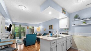 A modern kitchen with white cabinets, a central island, and a dining area with a blue sofa. - Pretty Peacock: 2BR/2BA near SDC & Lake Access! (Branson West)