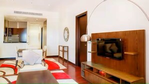 A modern living room with a flat-screen TV mounted on a wooden wall, a white sofa, a red and white rug, and a dining area visible through an open door. - Luxury 1- Bedroom at Anantara Palm Jumeirah (Dubai)