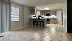 A modern kitchen with grey cabinets, a central island with two chairs, a ceiling fan, and a large window with blinds. - Multi Family House 4Bed/3Bath-Large Fenced Yard with Activities for Whole Family (Fruitland Park)