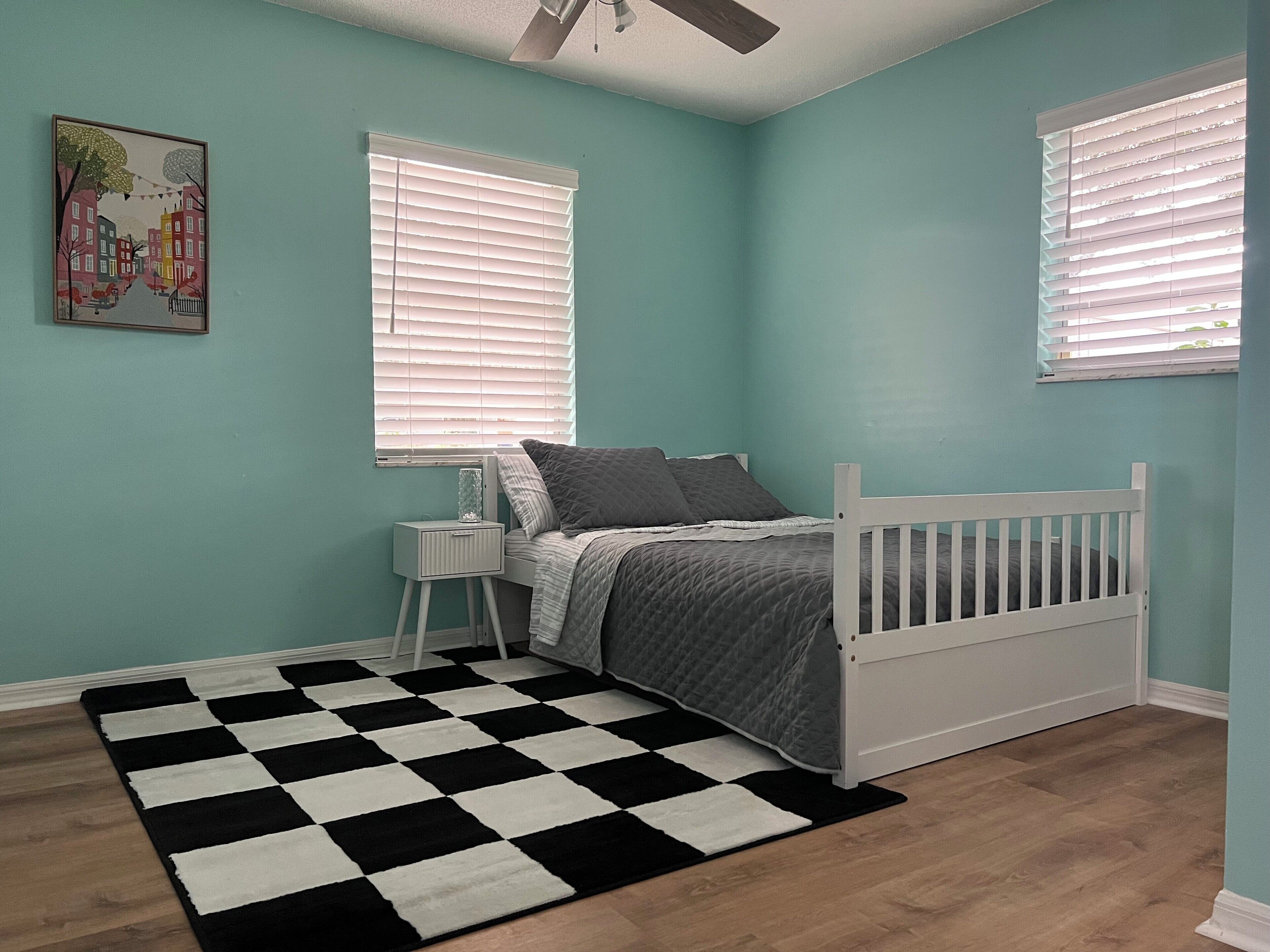 Quarto com paredes verde-água, estrutura de cama branca, tapete quadriculado preto e branco, e duas janelas com persianas.