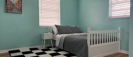 Quarto com paredes verde-água, estrutura de cama branca, tapete quadriculado preto e branco, e duas janelas com persianas.