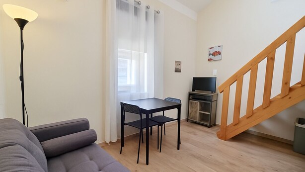 A compact living space with a sofa, a dining table with chairs, a TV on a cabinet, and a wooden staircase.