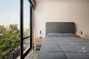 Ein Schlafzimmer mit großem Fenster, einem Bett mit grauem Kopfteil, einem Nachttisch mit Lampe und Blick auf eine Stadtansicht.