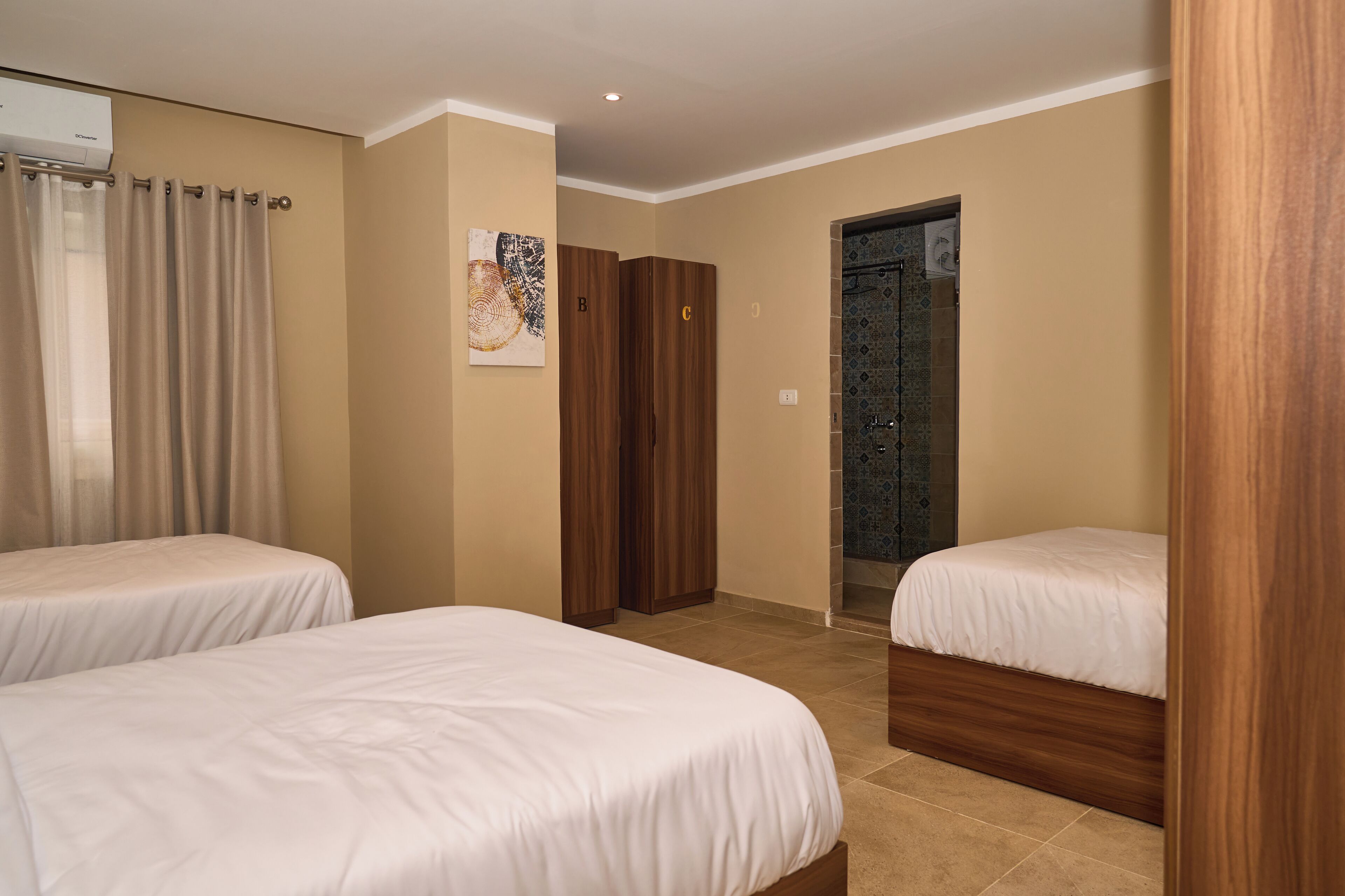 A hotel room with two single beds, a wooden wardrobe, a bathroom with a shower, and a wall-mounted air conditioner.