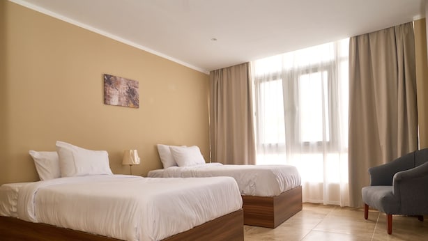 A hotel room with two beds, a chair, a painting on the wall, and large windows with curtains. - The Fit Stop Hostel (Hurghada)