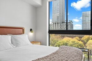 A hotel room with a bed, a nightstand, a lamp, and a large window with a city view. - Miami Luxury Condo (Miami)